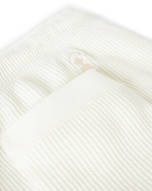 A close-up of The Cannes Waffle Knit Shorts - Vintage Ivory by Dandy Del Mar, with a small embroidered logo and a rectangular tag sewn onto it.