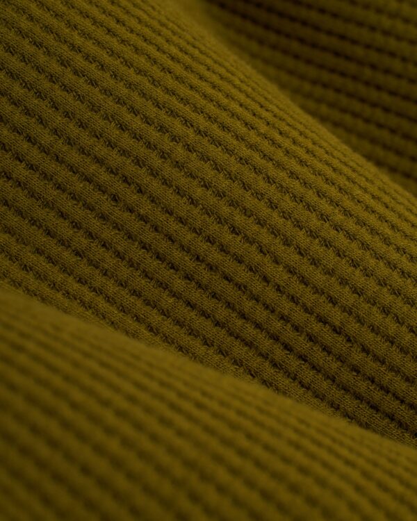 Discover the intricate beauty of The Cannes Waffle Knit Tee - Arbequina by Dandy Del Mar, featuring a mustard yellow color that mimics sophisticated waffle-knit textures with a repeating square pattern and inviting warmth.
