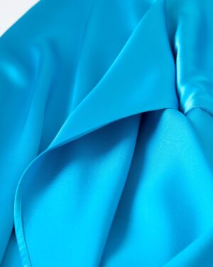Close-up of The Catania Satin Angel Sleeve Dress - Lago by Dandy Del Mar, showcasing its vibrant blue satin fabric with smooth folds and a glossy finish.