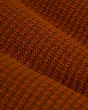 Close-up of The Sebastian Long Sleeve Polo in Burnt Sienna by Dandy Del Mar, showcasing a brown textured knit fabric with a repeating wavy pattern, reminiscent of an intricate design.
